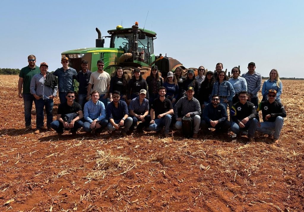 Grupo de estudantes de Agronomia da Universidade Estadual de Maringá (UEM) posando em frente a um trator na Fazenda Hervalense em Diamantino, Mato Grosso, durante visita técnica em 10 de agosto de 2024. Ao fundo, uma área de plantação sob céu azul.
