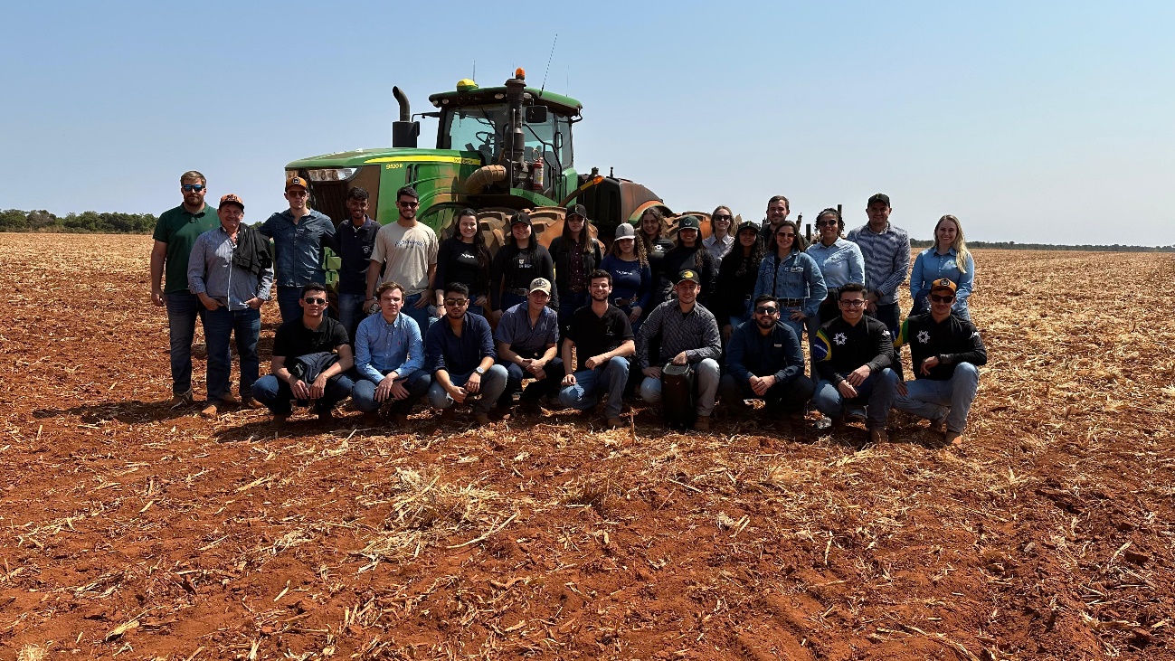 Grupo de estudantes de Agronomia da Universidade Estadual de Maringá (UEM) posando em frente a um trator na Fazenda Hervalense em Diamantino, Mato Grosso, durante visita técnica em 10 de agosto de 2024. Ao fundo, uma área de plantação sob céu azul.