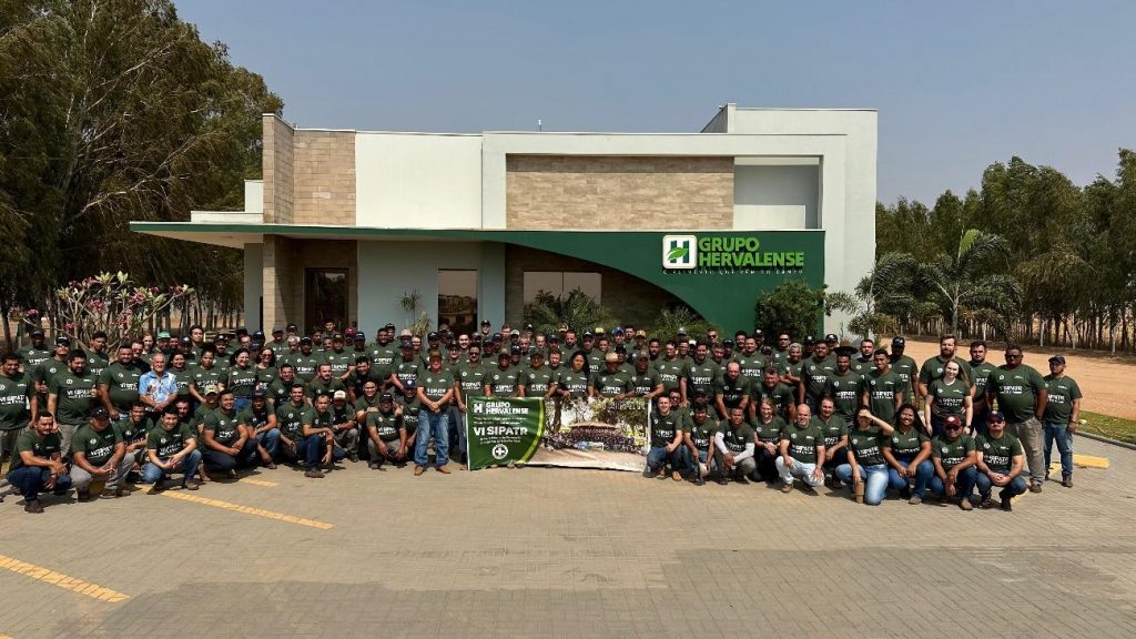 Uma grande foto de grupo com dezenas de colaboradores do Grupo Hervalense, todos usando camisetas verdes da 6ª SIPATR, posando em frente ao edifício da empresa. A imagem destaca o comprometimento da equipe com a segurança no ambiente de trabalho rural.