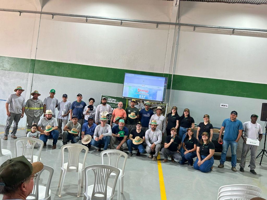 Um grande grupo de colaboradores, vestindo roupas de trabalho e chapéus de palha, posa sorrindo em um galpão espaçoso. Alguns seguram brindes recebidos durante o evento, com uma tela ao fundo exibindo uma mensagem de incentivo à participação.