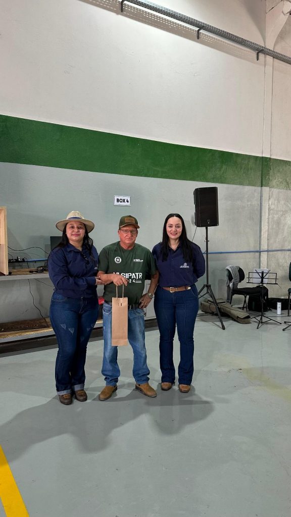 Três pessoas estão posicionadas em frente a uma parede de cor verde e branca no galpão, uma delas segurando um brinde. Todos estão sorrindo e vestindo uniformes da empresa, demonstrando o clima amistoso do evento.