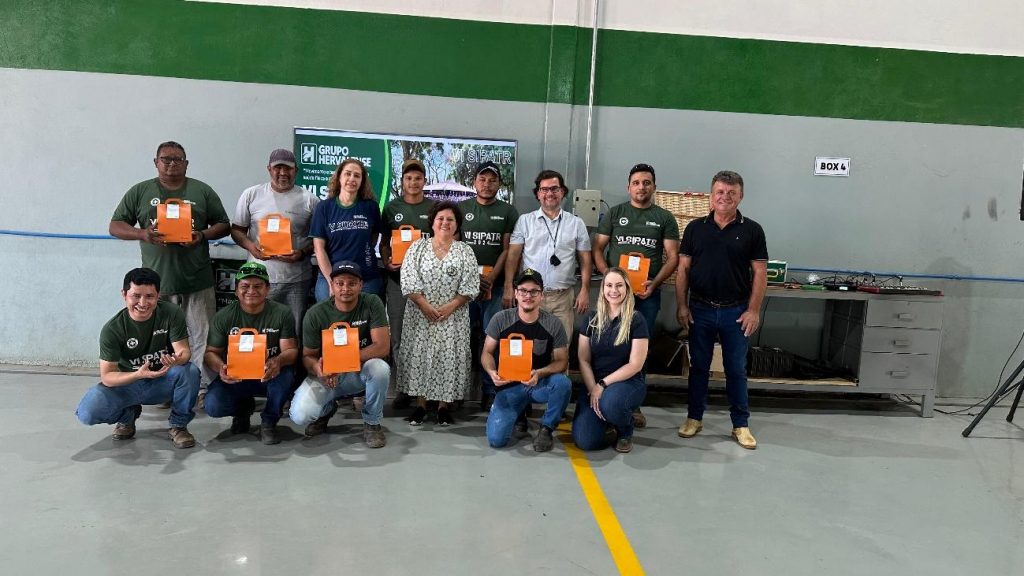 Grupo de colaboradores do Grupo Hervalense, composto por homens e mulheres, posa sorridente, segurando sacolas laranjas. O cenário inclui um ambiente de trabalho limpo e organizado, demonstrando o sucesso do evento e o reconhecimento dos participantes.
