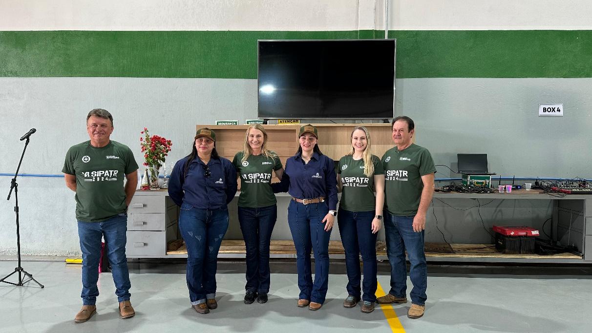 Seis membros da equipe do Grupo Hervalense, usando camisetas verdes com a inscrição "VI SIPATR 2024", estão alinhados em frente a uma bancada com uma televisão ao fundo. Eles sorriem e parecem satisfeitos, reforçando o espírito de união do evento.