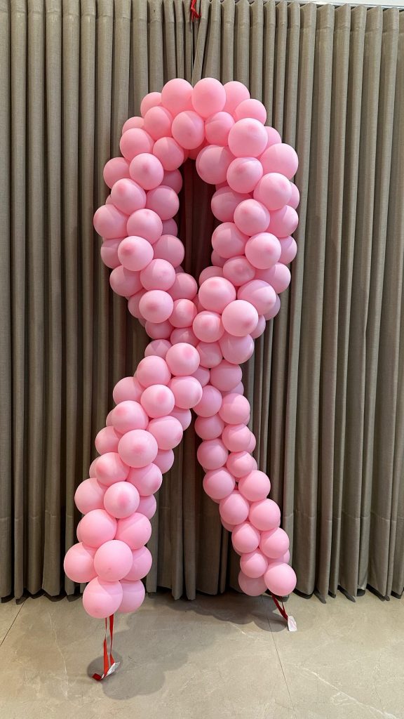 Um laço rosa grande feito de balões, simbolizando a campanha de conscientização sobre o câncer de mama. O laço está pendurado na parede, em frente a uma cortina de cor neutra. A decoração é simples e delicada, destacando a causa do Outubro Rosa e a prevenção ao câncer.