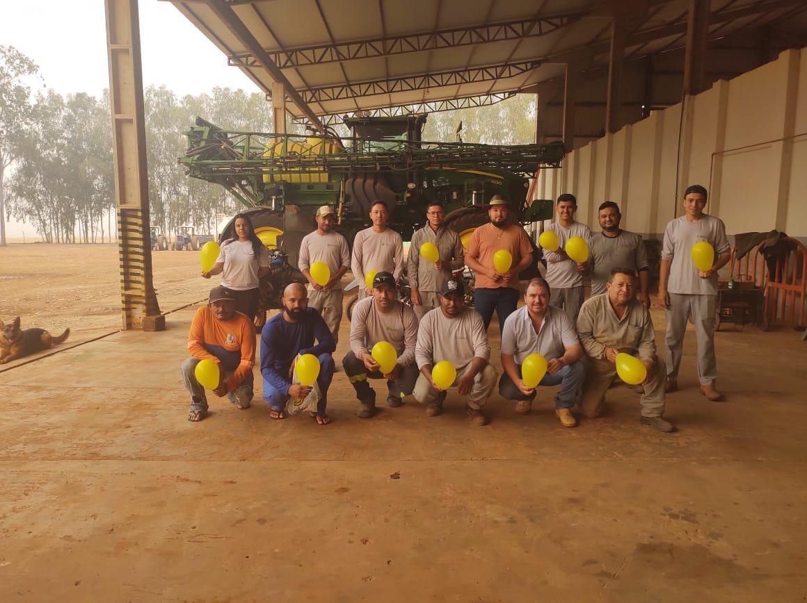 Colaboradores do Grupo Hervalense reunidos em um ambiente coberto, com máquinas agrícolas ao fundo. Todos estão segurando balões amarelos, em um gesto de apoio à Campanha Setembro Amarelo, com foco na prevenção ao suicídio e no cuidado com a saúde mental.