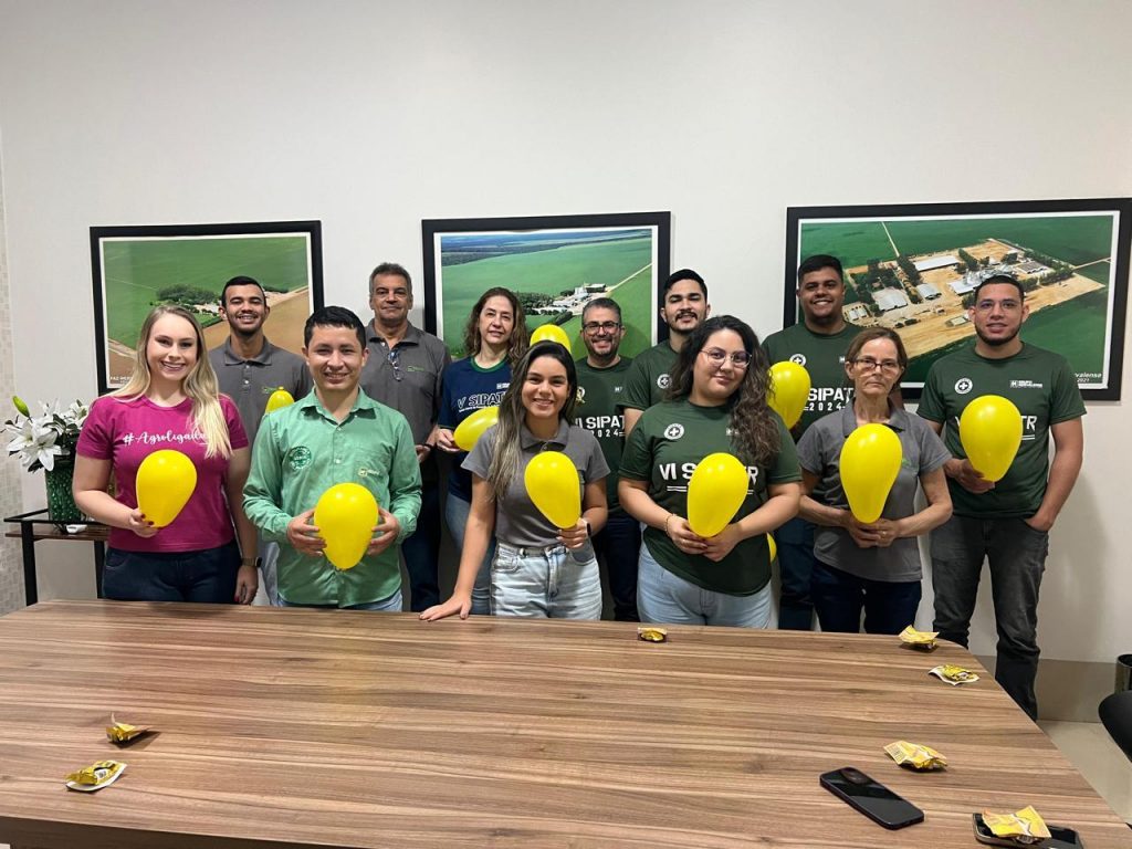 Um grupo diverso de colaboradores do Grupo Hervalense, dentro de uma sala de reuniões, segurando balões amarelos. A imagem captura a participação interna na Campanha Setembro Amarelo, ressaltando a união dos times em prol da conscientização sobre saúde mental.