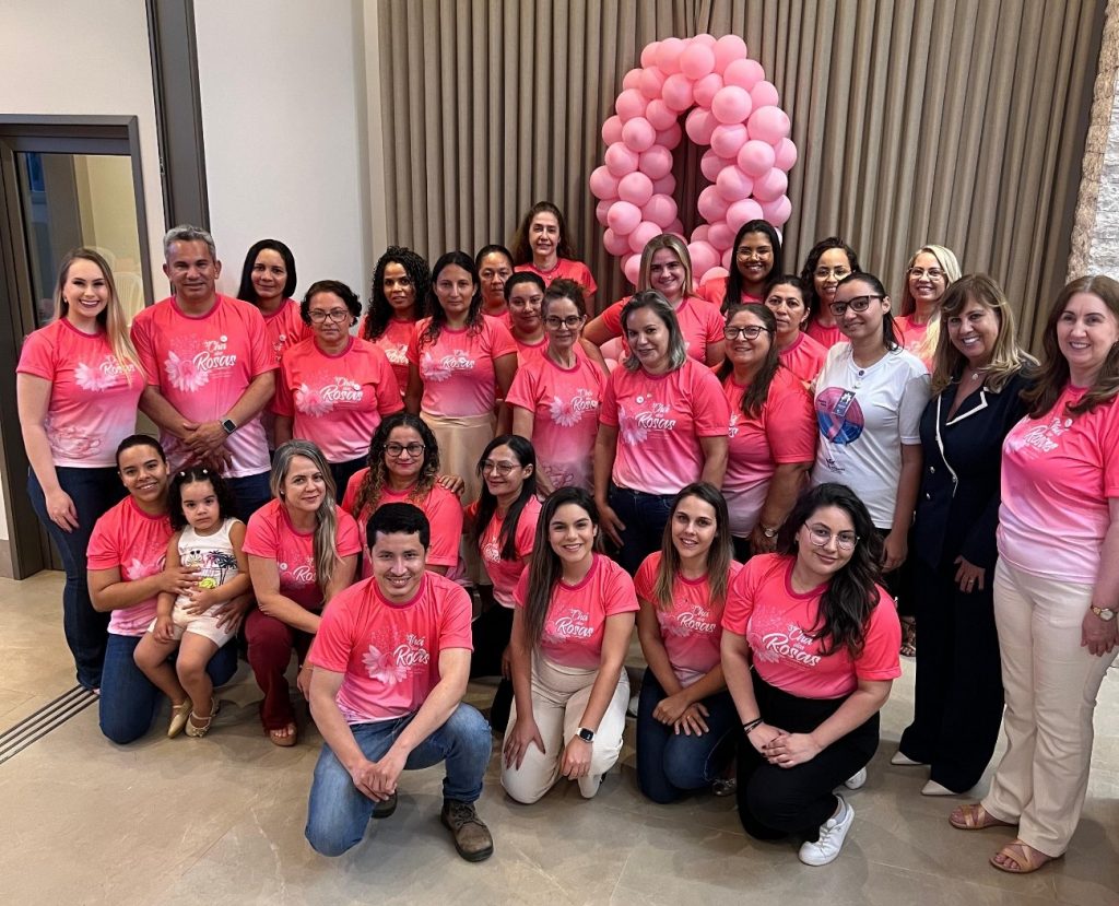 Foto em grupo do evento 3º Chá das Rosas, com várias mulheres vestindo camisetas cor-de-rosa e posando em frente a um grande laço rosa feito de balões, símbolo da campanha Outubro Rosa. O grupo, composto por colaboradores do Grupo Hervalense, inclui mulheres de diferentes idades e um homem, todos participando da conscientização sobre a prevenção do câncer de mama e colo do útero.