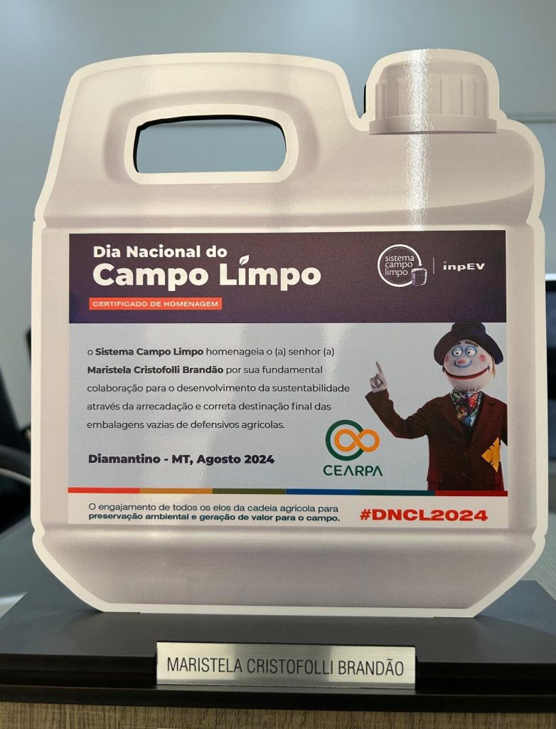 Placa de homenagem no formato de galão branco com texto impresso destacando o "Dia Nacional do Campo Limpo". O texto informa que o Sistema Campo Limpo homenageia Maristela Cristofolli Brandão por sua contribuição à sustentabilidade por meio da arrecadação e destinação correta de embalagens vazias de defensivos agrícolas. A homenagem foi concedida em Diamantino - MT, em agosto de 2024. Inclui os logotipos do Sistema Campo Limpo, inpEV e CEARPA, além da hashtag #DNCL2024. No canto direito, uma ilustração de um mascote sorridente e vestindo um terno aponta para o texto. Na base da placa, há uma etiqueta metálica com o nome "Maristela Cristofolli Brandão"