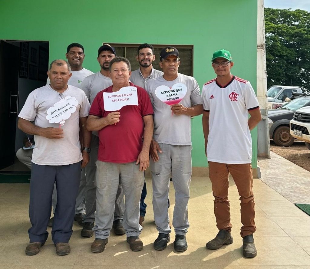 Colaboradores do Grupo Hervalense posam sorridentes em frente à fachada verde da Fazenda Hervalense, segurando placas com frases incentivando a doação de sangue.