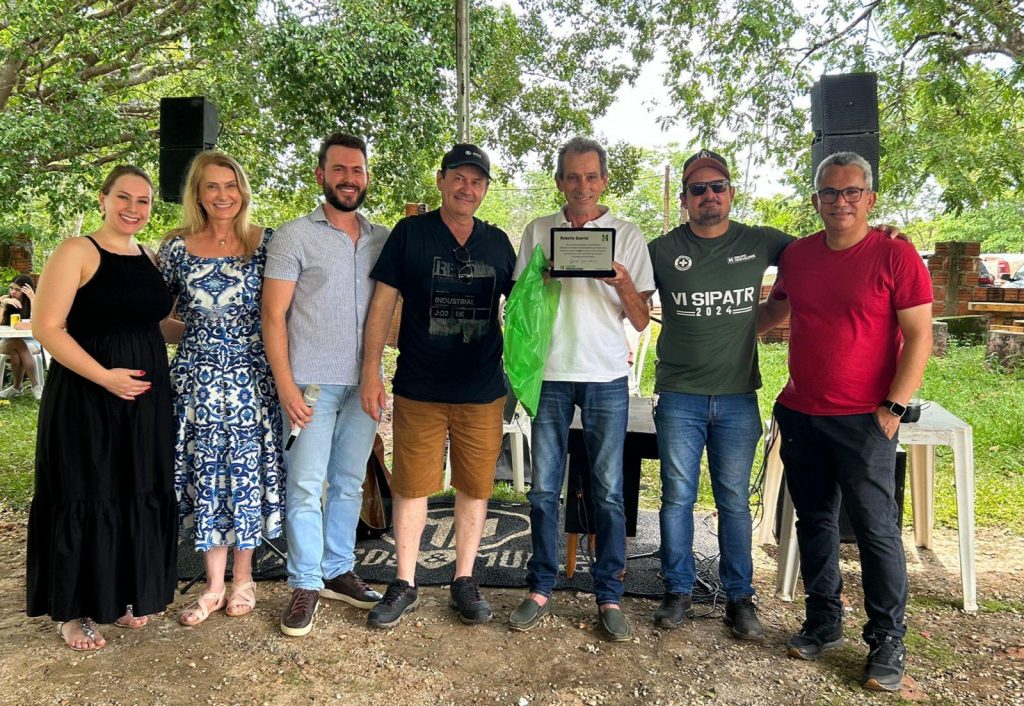 Foto final com a equipe do Grupo Hervalense reunida em frente ao palco, sorrindo e segurando uma placa de homenagem. Um colaborador ao centro é celebrado, simbolizando reconhecimento e união entre colegas.