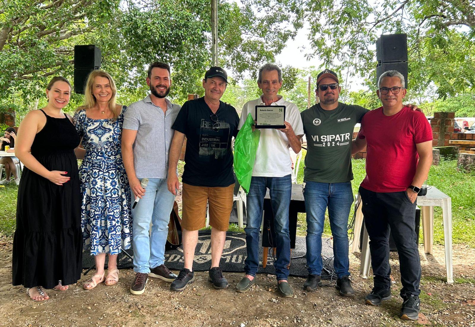 Foto final com a equipe do Grupo Hervalense reunida em frente ao palco, sorrindo e segurando uma placa de homenagem. Um colaborador ao centro é celebrado, simbolizando reconhecimento e união entre colegas.