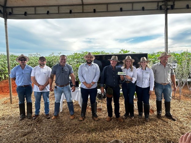 Oito representantes do evento posando para foto sob uma cobertura no campo, com lavouras de mamona ao fundo e um monitor sobre mesa atrás do grupo.