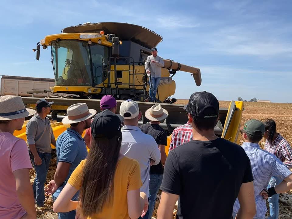 Grupo de pessoas participando de demonstração técnica sobre agricultura digital, com especialista explicando o uso de máquina agrícola avançada em campo.