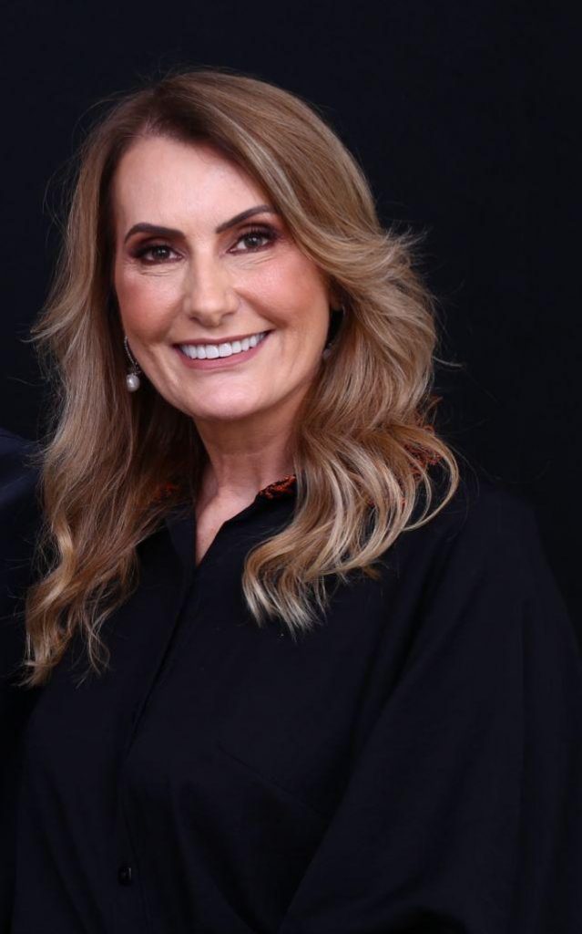 Retrato de Marinês Christofolli Parisenti, sócia-fundadora e gestora do Grupo Hervalense. Ela aparece sorridente, com maquiagem leve e cabelos castanhos claros soltos, usando blusa preta com detalhes discretos. O fundo escuro valoriza a expressão confiante e acolhedora, transmitindo a imagem de liderança feminina, resiliência e inovação no agronegócio.