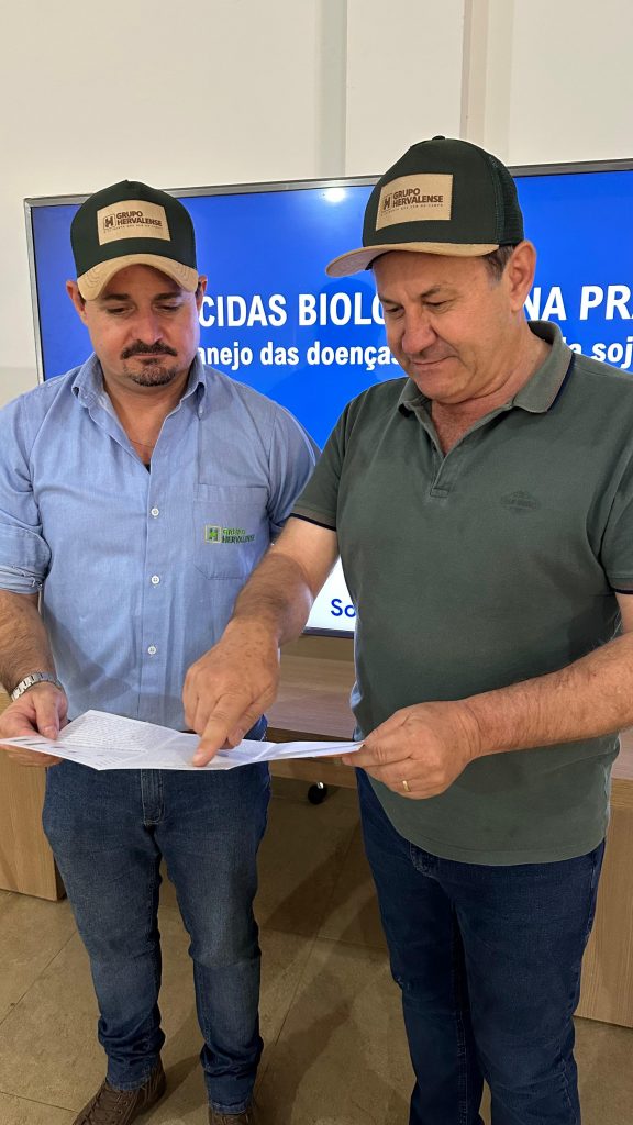 Registro em ambiente interno do Grupo Hervalense. Victor Sanches, à esquerda, de camisa azul clara com logomarca bordada, e Erny Parisenti, à direita, de camisa verde escura e boné, analisam juntos documentos impressos. Ambos aparecem concentrados, em frente a um telão de apresentação que exibe conteúdo sobre manejo de doenças na soja e uso de defensivos biológicos. A cena destaca a combinação de conhecimento técnico e tomada de decisão estratégica em prol da sustentabilidade ambiental e da inovação agrícola.