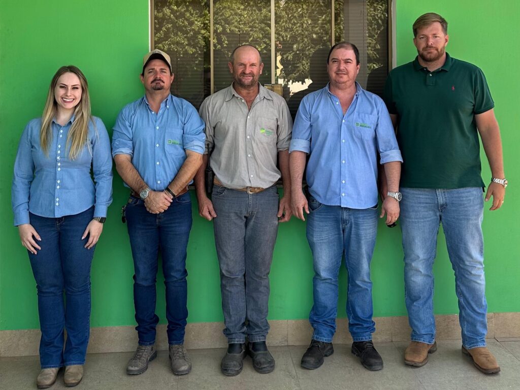 Foto em grupo de cinco profissionais do Grupo Hervalense, posando lado a lado em frente a um prédio com fachada verde e logotipo em destaque na parte superior. Todos estão vestidos com camisas sociais em tons de azul, cinza e verde, representando a equipe técnica da empresa. A imagem transmite união, profissionalismo e valorização da agronomia no contexto do Dia do Agrônomo.